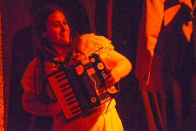 foto de uma apresentação de teatro. Uma jovem branca, de cabelos longos e castanhos, vestida de branco, segurando um acordeon. Uma luz avermelhada ilumina a jovem atriz.