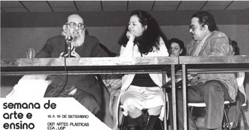 Foto em preto e branco de palestrantes na Semana de Arte e Ensino. Na esquerda, Paulo Freire, homem branco, calvo, com cabelos nas laterais da cabeça e barba comprida e grisalha. Usa um óculos de aro preto quadrado e terno escuro. Está falando em um microfone. Ao lado dele e no centro da foto, Ana Mae Barbosa, mulher branca com cabelos longos, pretos e ondulados, veste uma blusa preta, um blazer e saia brancos e botas pretas. Ela olha para Paulo Freire com o rosto apoiado na mão direita. Ao lado dela e à direita da foto, o professor Alexandre Barbosa, homem branco de cabelos pretos e bigode. Ele veste um terno xadrez, calça social escura e óculos de armação preta. Todos se sentam atrás de uma mesa.