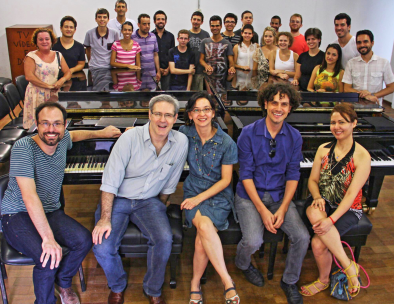 Grupo de pessoas posando para uma foto. Cinco pessoas, tres homens e duas mulheres, estão sentados em frente a dois pianos. Atrás dos instrumentos, um grupo de cerca de 20 pessoas, uma ao lado da outra, está de pé. Todos olham para a câmera e sorriem.
