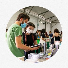 um homem branco, de cabelos curtos, lisos e castanhos e barba da mesma cor, vestindo uma camisa verde, olha para baixo, segurando um livro que tem em suas mãos. Ele mostra o livro para uma jovem branca, de cabelos longos e castanhos até a metade e rosa nas pontas, presos em duas tranças. Ambos usam mascaras sobre o rosto. Eles estão em frente de uma mesa, com vários livros expostos. Ao fundo, pessoas circulam entre outras mesas com livros expostos