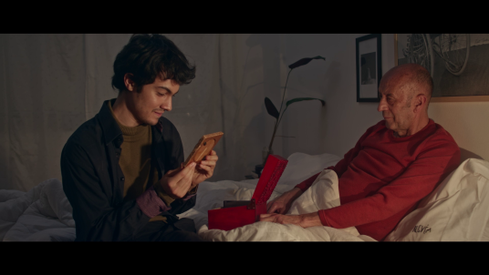 Cena do curta-metragem Olhares Trêmulos. Um homem de idade, pele branca e sem cabelos, está sentado em uma cama com as pernas cobertas por lençóis brancos. Ele veste uma camiseta vermelha de mangas compridas. Ao lado dele, em cima da cama, uma caixa vermelha está aberta. À sua frente, sentado na cama, um rapaz de pele branca, cabelos escuros e lisos olha para um objeto e sorri levemente. O rapaz veste uma camisa de botões escura sobre uma camiseta marrom.