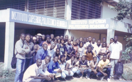 Foto de estudantes em frente ao prédio do Instituto. Diversos homens e mulheres estão reunidos posando para a foto. Alguns estão em pé e outros estão abaixados. Todos são negros. A fachada do prédio é branca e tem algumas janelas. Duas vigas da fachada estão pintadas com os dizeres  “Instituto Superior Politécnico Benhoblô”, em letras brancas com fundo azul 