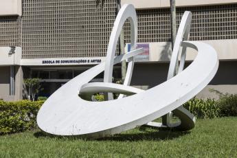 Foto de parte da fachada do prédio da ECA. Sobre um gramado verde, está uma escultura branca formada por quatro arcos que se entrelaçam entre si: um à frente, dois paralelos um ao outro e um quarto arco, mais próximo ao chão. Atrás da escultura, parte da fachado do prédio. A fachada tem elementos vazados, do tipo cobogó, na cor bege. O nome da escola está afixado na parede, com as letras na cor azul escuro.