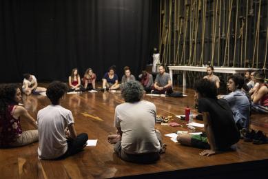 Grupos de estudantes estão sentados, em forma de circulo, no palco de um teatro.