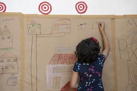 Foto de uma menina de pele clara e cabelos escuros que desenha com giz de cera em um papel kraft colado na parede. Ela está de costas e escreve ‘museu’ em um retângulo desenhado ao lado de outros prédios e casas, ligados por ruas. No centro do desenho há uma grande casa com telhado vermelho e paredes rosa claro.