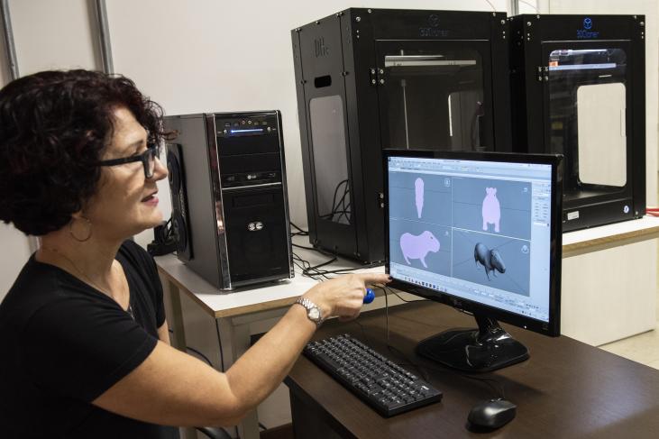 Monica Tavares é uma mulher branca, cabelos pretos, médios e ondulados e usa óculos. Ela aponta para a tela de um computador com a imagem de um animal em diferentes ângulos, preparando para impressão 3d