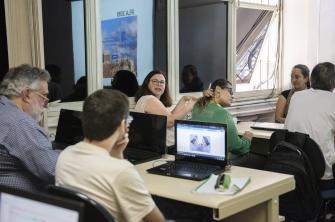 Foto de pessoas em uma sala de aula: homens e mulheres de diferentes faixas etárias, todos brancos. Eles estão sentados diante de mesas e a maioria têm notebooks à sua frente. Ao centro, uma mulher branca, de cabelos castanhos escuros, óculos redondos e blusa de cor clara fala e gesticula com o corpo voltado para as pessoas sentadas atrás dela. Alguns a observam. Ao fundo, janelas refletem uma tela onde está sendo projetada a fotografia de uma paisagem urbana. 