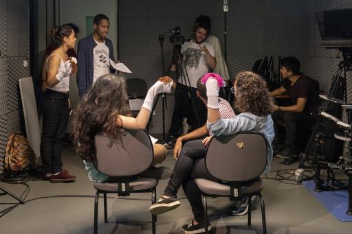 Oito estudantes estão reunidos em um estudio de gravação. Duas jovens estão de costas, sentadas cada uma em uma cadeira, e de frente uma para a outra. Elas tem em uma das mãos um fantoche. Um jovem está agachado na frente delas. Os três observam, ao fundo, um jovem homem de camiseta branca, que manipula uma camera em um tripé. Do lado esquerdo, estão, de pé, um jovem, segurando uma folha de papel, e uma jovem, também com um fantoche em uma das mãos. À direita, um aluno está sentado em uma cadeira.