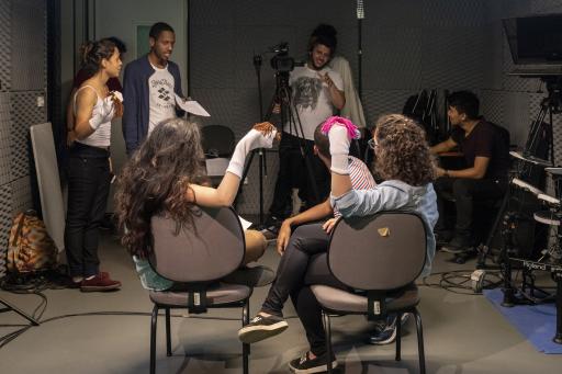 Oito estudantes estão reunidos em um estudio de gravação. Duas jovens estão de costas, sentadas cada uma em uma cadeira, e de frente uma para a outra. Elas tem em uma das mãos um fantoche. Um jovem está agachado na frente delas. Os três observam, ao fundo, um jovem homem de camiseta branca, que manipula uma camera em um tripé. Do lado esquerdo, estão, de pé, um jovem, segurando uma folha de papel, e uma jovem, também com um fantoche em uma das mãos. À direita, um aluno está sentado em uma cadeira. 