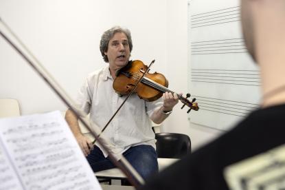 Foto de um homem branco, de cabelos grisalhos, sentado e tocando uma viola erudita. Ele veste uma camisa clara de mangas curtas e calça escura. De frente para ele está uma pessoa de costas para a foto, ao lado de uma partitura. A sala é branca e há uma lousa de vidro com linhas de partitura.