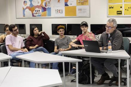 Foto de um professor e estudantes durante aula. À direita, sentado em uma mesa, o professor, um homem branco de cabelos grisalhos, óculos, calça jeans e camisa cinza, fala e gesticula com um laptop à sua frente. Sentados em cadeiras a seu lado, um grupo de jovens o escuta e observa. Alguns sorriem. Ao fundo, diversos computadores e paredes com murais cobertos por cartazes coloridos, aparentemente produções gráficas estudantis. 