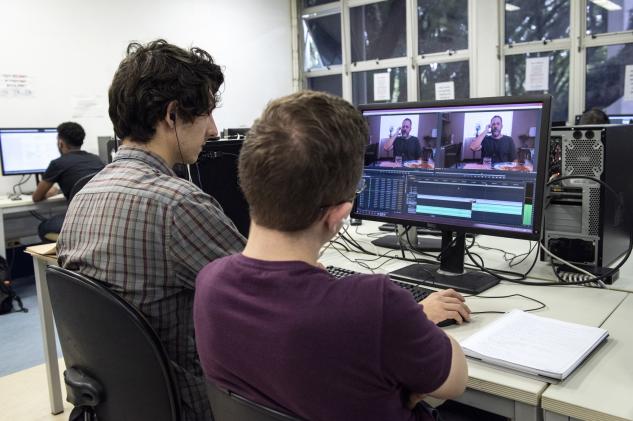 Alunos de costas editando vídeos no computador do laboratório.