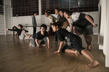 Foto de diversas pessoas fazendo uma performance em um estúdio. Cada um faz uma pose diferente, mas olham na mesma direção. A maioria veste roupas escuras.