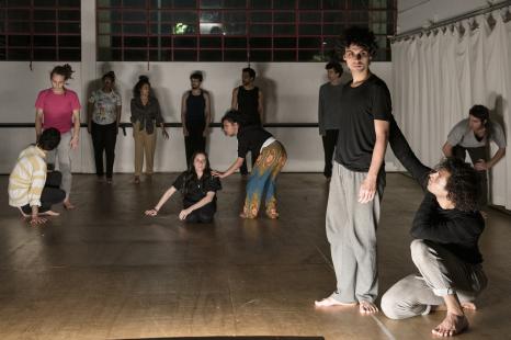 Foto. Em uma sala ampla com piso de madeira, paredes brancas, vitrô ao fundo e uma cortina branca no canto direito, um grupo de pessoas participa de uma aula de dança e expressão corporal. À frente, estão dois homens, jovens, brancos, com cabelos castanhos e ondulados; um está de pé e olha para baixo com expressão séria, o outro está agachado, erguendo um dos braços e tocando nas costas do primeiro. Eles se vestem de forma semelhante: calça cinza e blusa preta, descalços. Atrás deles estão outras cinco pessoas: à esquerda, dois homens interagem, um de pé e outro agachado; ao centro, uma mulher de pé, curvada, toca o ombro de outra, que está sentada; à direita, um homem está curvado, com as mãos sobre os joelhos, observando as mulheres que estão próximas a ele. Ao fundo, outras cinco pessoas estão de pé, em linha próxima à parede, olhando uns para os outros.