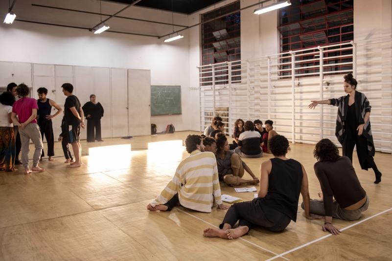 foto de cerca de 20  jovens em uma sala ampla. São, na maioria, brancos, e dividem-se em três grupos. Dois grupos estão sentados no piso e outro está de pé. Todos estão descalços e usam roupas largas e confortáveis. Entre os dois grupos sentados, destaca-se uma mulher jovem que fala e gesticula. A sala tem piso de madeira, duas grandes janelas e barras de alongamento. No fundo da sala há uma lousa verde e um grande armário, à frente do qual uma mulher está de pé observando os grupos. 