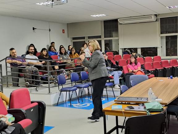 Foto em que uma mulher de pele branca e cabelo claro está em pé em um auditório. Ela fala enquanto outras pessoas, homens e mulheres, estão sentados em poltronas a olhando. Do lado direito do auditório algumas poltronas estão vazias. Atrás da palestrante há uma mesa de madeira. Do seu lado esquerdo existem mais pessoas também sentadas, mas não aparecerem na fotografia.