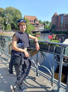 Fotografia de uma pessoa apoiando um dos cotovelos sobre a grade de uma ponte às margens de um lago. Ela tem pele clara, cabelos claros e curtos e traja roupas pretas: camiseta regata, calça, tênis, casaco amarrado na cintura, gorro, bolsa. Há flores em uma jardineira presa à grade e, do outro lado do lago, há árvores e três casarões avermelhados.