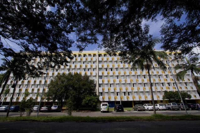 Foto de um prédio amarelo retangular extenso, com muitas janelas. Ele está parcialmente coberto por copas de árvores no primeiro plano da imagem. No térreo estão alguns carros estacionados e parte do estacionamento e da calçada.