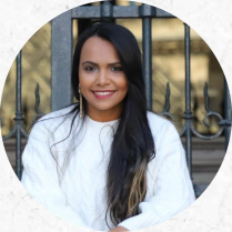 Foto de uma mulher jovem, parda de olhos castanhos, cabelos lisos, longos e pretos com as pontas descoloridas. Ela usa blusa branca de mangas longas e brincos grandes dourados. Ela sorri e atrás dela há um portão de ferro.