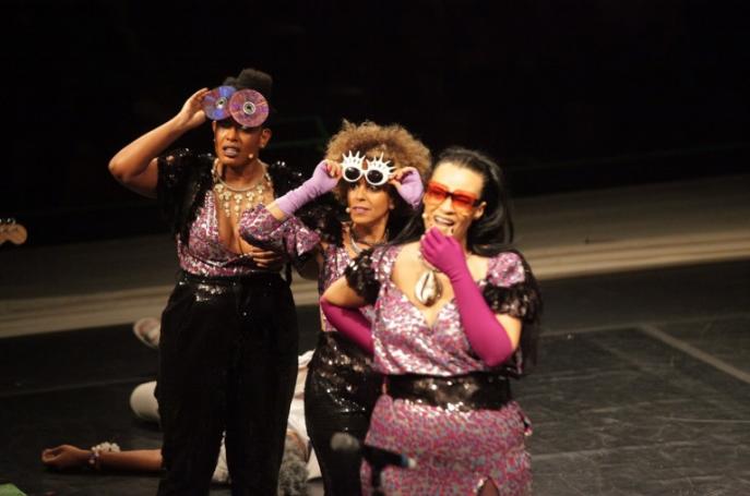 Fotografia colorida de três atrizes negras olhando para algo fora do palco. As três utilizam roupas em cores rosa e preto metálicos e óculos personalizados com CDs. Atrás delas, caída sobre o palco, está uma quarta atriz, de cabelos grisalhos e vestida com roupas brancas. 