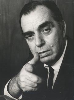 Foto em preto e branco. Nelson Rodrigues, um homem branco, de cabelo preto, que usa um terno. Aponta com a mão "em formato de arma" para a câmera.