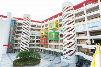 Foto da fachada da universidade. O prédio apresenta uma estrutura moderna, com três torres brancas espiraladas e alguns cubos coloridos projetados para fora da fachada. No canto inferior esquerdo da imagem, encontra-se uma parte elevada, na qual há um canteiro grande e florido.  