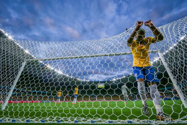 Foto de um jogador da Seleção Brasileira se agarrando de cabeça baixa na rede de um gol. Ao fundo, encontram-se mais três jogadores, incluindo o goleiro. A foto foi tirada de baixo para cima, de modo que o céu ocupa a metade superior da imagem, emoldurado pelas bordas do estádio Mineirão.