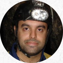 Foto de rosto de um homem branco, de olhos castanhos e barba por fazer. Ele usa capacete preto com lanterna pequena acoplada e camisa azul e amarela. Atrás dele há uma parede rochosa.