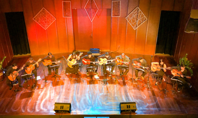 Fotografia a partir do alto de um palco de madeira no qual estão dez violonistas sentados em cadeiras dispostas em um semicírculo. O palco é iluminado por uma mistura de tons quentes e frios. Os músicos trajam roupas escuras e tocam seus violões enquanto olham partituras dispostas diante de cada um. Nas laterais do palco há vasos de plantas e, ao fundo, duas coxias com cortinas. O cenário é composto por painéis de madeira e, no centro, há uma porta dupla e quatro quadros decorativos com linhas criando formatos geométricos.