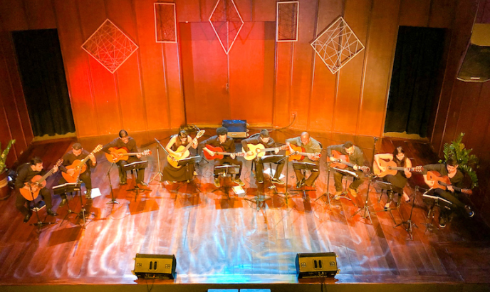 Fotografia a partir do alto de um palco de madeira no qual estão dez violonistas sentados em cadeiras dispostas em um semicírculo. O palco é iluminado por uma mistura de tons quentes e frios. Os músicos trajam roupas escuras e tocam seus violões enquanto olham partituras dispostas diante de cada um. Nas laterais do palco há vasos de plantas e, ao fundo, duas coxias com cortinas. O cenário é composto por painéis de madeira e, no centro, há uma porta dupla e quatro quadros decorativos com linhas criando formatos geométricos. 