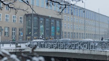 foto da fachada da universidade, num primeiro plano, uma ponte de metal e concreto, com letras metálicas escritas Universitat zu Koln. Carros passam pela ponte. Ao fundo, um prédio retangular marrom com janelas na cor branca. A estatua de um homem pensando está a frente do prédio. Está nevando.