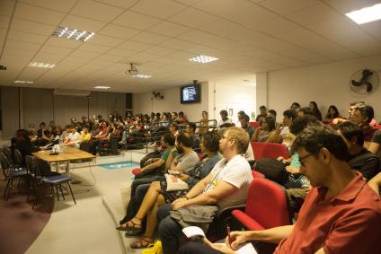 Fotografia do auditório sede da 5ª Jornada. Na imagem pessoas estão sentadas nos dois lados do auditório enquanto olham para uma apresentação que não aparece na foto. A maioria das cadeiras está ocupada.