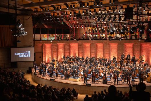 Foto de um auditório com a platéia cheia, assim como o palco, com uma orquestra sinfônica que constitui o foco da imagem. Os integrantes da orquestra estão de pé, recebendo os aplausos dos ouvintes. No teto, encontram-se pendurados dezenas de holofotes, os quais dão ao palco uma iluminação amarelada. Na porção esquerda da imagem, uma tela exibe o logo do Festival de Inverno de Campos do Jordão.