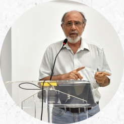 Foto de um homem idoso de pele branca com barba, bigode e cabelos grisalhos. Ele utiliza óculos redondos de armação fina, camisa social branca, calça jeans, cinto marrom e relógio. Ele está diante de um púlpito em acrílico transparente e fala ao microfone enquanto aponta para o lado direito da imagem com ambas as mãos.