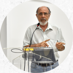 Foto de um homem idoso de pele branca com barba, bigode e cabelos grisalhos. Ele utiliza óculos redondos de armação fina, camisa social branca, calça jeans, cinto marrom e relógio. Ele está diante de um púlpito em acrílico transparente e fala ao microfone enquanto aponta para o lado direito da imagem com ambas as mãos. 