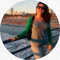 Foto de uma mulher branca, de cabelos ondulados, castanhos e na altura dos ombros. Ela sorri e usa óculos de sol, camiseta cinza, calça jeans de lavagem clara, casaco verde e leva bolsa preta nos ombros. Atrás dela há um guarda corpo de ferro e uma praia