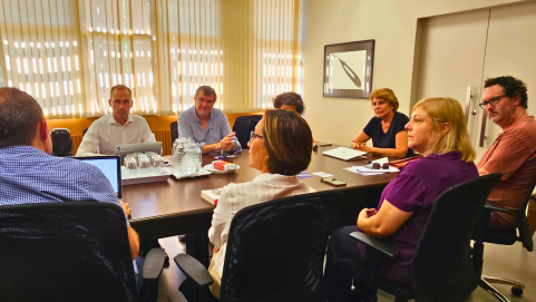 Foto de um grupo de pessoas sentado ao redor de uma mesa retangular. Na frente, de costas ou de lado para a foto, estão os docentes da ECA: dois homens e duas mulheres, sentados em cadeiras de escritório, na cor preta. Ao fundo, e de frente para a foto, estão três docentes da faculdade francesa. Na cabeceira da mesa, está sentada a diretora da ECA. Em cima da mesa de madeira, na cor marrom escuro, estão diversos objetos, jarras de água, copos de vidro, computadores, papeis, livros, jarra de café. Atrás da mesa, persianas na cor bege cobrem as janelas, e, na parede de cor semelhante, está pendurado um quadro.