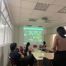 Foto colorida em ambiente interno, com pessoas ao redor de uma mesa vendo uma projeção digital na parede