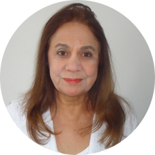 Foto do rosto de uma mulher idosa, de pele clara, de olhos castanhos e cabelos castanhos longos, lisos e compridos. Ela olha para a câmera, sorri levemente e usa uma blusa branca. Atrás dela, uma parede branca.