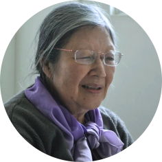 Foto de uma mulher idosa com traços amarelos. Os cabelos longos, lisos e grisalhos estão presos. Ela usa óculos de lentes retangulares, blusa cinza e lenço lilás. Atrás dela, uma parede branca.
