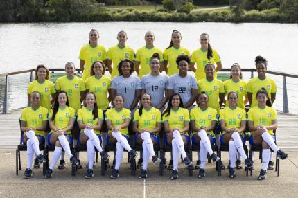 Foto da Seleção Brasileira Feminina principal de 2023. 23 mulheres posam para uma foto, sendo 14 em pé e, à frente delas, nove sentadas em cadeiras com as pernas cruzadas. Três das mulheres, que estão em pé e no centro da fotografia, usam uniformes cinzas, as demais usam uniformes amarelo e azul e meião branco. Elas estão no que aparenta ser um cais, todas sorriem para a câmera.