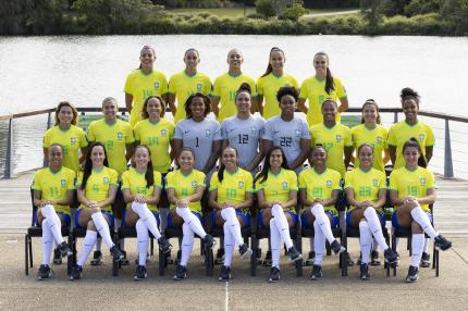 Foto da Seleção Brasileira Feminina principal de 2023. 23 mulheres posam para uma foto, sendo 14 em pé e, à frente delas, nove sentadas em cadeiras com as pernas cruzadas. Três das mulheres, que estão em pé e no centro da fotografia, usam uniformes cinzas, as demais usam uniformes amarelo e azul e meião branco. Elas estão no que aparenta ser um cais, todas sorriem para a câmera. 