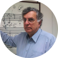 Foto de um homem branco, de cabelos curtos, ondulados e grisalhos. Ele usa óculos retangulares e camisa azul e olha diretamente para a câmera, com expressão séria. Atrás dele, uma lousa branca, com linhas pretas, tal qual uma partitura de música, repleta de anotações.