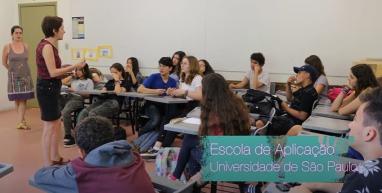 Print de vídeo do canal do Foca nas Mídias. Na imagem, há vários estudantes sentados em uma sala de aula. Todos estão voltados para uma mulher branca, de cabelo escuro e curto, que está em pé e parece explicar algo. Ao fundo, perto de uma porta amarela, está outra mulher, também olhando para a moça que está na frente dos alunos.