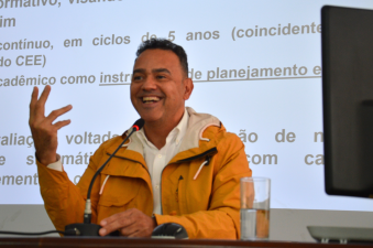 Homem branco, de cabelos castanhos e curtos, vestindo uma camisa branca e uma jaqueta amarela, está sentado e falando ao microfone. Ao fundo, a projeção de slide com letras na cor preta sob um fundo branco.