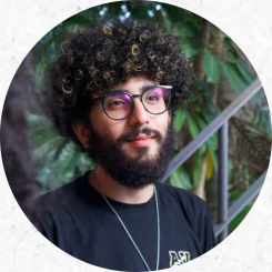 retrato de um homem branco de barba escura. Seu cabelo é cacheado e preto com mechas loiras na parte da frente. Ele usa uma camiseta preta com um logo pequeno de contorno amarelo na altura do peito, uma corrente prateada comprida e fina e um óculos de armação também preta e formato arredondado. O recorte da foto vai até a altura do peito e ele não olha diretamente para a câmara, estando virado ligeiramente para a diagonal direita da imagem. Atrás dele há um fundo verde composto por plantas e uma estrutura de ferro que se assemelha a um corrimão de uma escada.