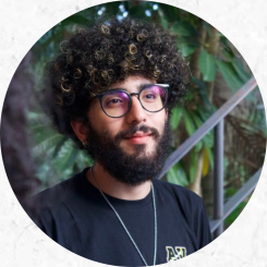 retrato de um homem branco de barba escura. Seu cabelo é cacheado e preto com mechas loiras na parte da frente. Ele usa uma camiseta preta com um logo pequeno de contorno amarelo na altura do peito, uma corrente prateada comprida e fina e um óculos de armação também preta e formato arredondado. O recorte da foto vai até a altura do peito e ele não olha diretamente para a câmara, estando virado ligeiramente para a diagonal direita da imagem. Atrás dele há um fundo verde composto por plantas e uma estrutura de ferro que se assemelha a um corrimão de uma escada. 