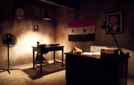 ​  Foto de uma sala mal iluminada e com paredes mofadas. Em primeiro plano e à direita, há uma mesa de escritório sobre a qual estão alguns papéis e livros. No fundo, há uma mesa mais simples, sobre a qual estão uma caixa e outro objeto. No fundo, mais à esquerda, há um ventilador de chão desligado. Nas paredes, há dois retratos de homem branco de cabelos curtos, e uma bandeira da Síria. 