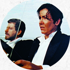 dois homens brancos, de cabelos curtos e castanhos, sentam-se lado a lado em um carro. Veste trajes formais