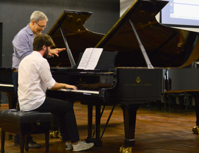 Foto de uma aula de piano. Um jovem, de cabelos castanhos e barba, está sentado de costas, com um piano preto a sua frente e tocando o instrumento. Ele veste uma camisa branca, calça preta e tenis. Ao seu lado, está um homem branco, de cabelos curtos e grisalhos, usando óculos e uma camisa azul e branca listrada. Ele observa as suas duas mãos, enquanto conversa com o jovem. O chão é de madeira e ao fundo uma parede preta e a vista parcial de um projetor